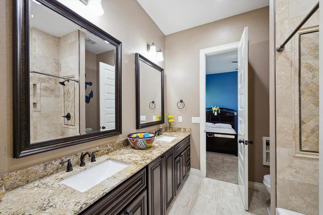 This bathroom features a dual-sink vanity with granite countertops and dark wood cabinets, complemented by large framed mirrors. It includes a tiled shower and a doorway leading to a bedroom, creating a seamless flow between spaces. | Image 12