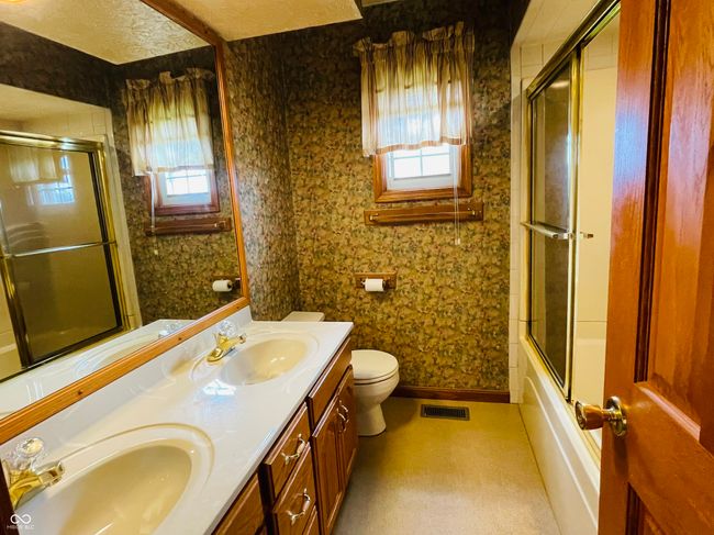 bathroom featuring toilet, double vanity, combined bath / shower with glass door, baseboards, and wallpapered walls | Image 16
