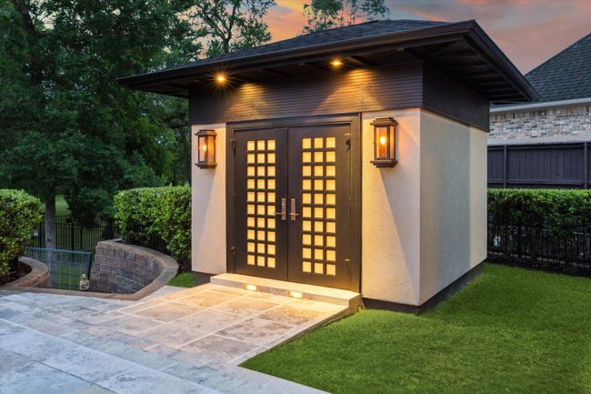This serene Zen-inspired pool house serves as a tranquil extension of the outdoor living space, thoughtfully designed to offer both form and function. Featuring a contemporary double-door entry flanked by warm gas sconces, it houses a convenient full bathroom and dressing room completing the resort-style ambiance of the backyard oasis. Also, note the stone stairway and iron gate leading to the creekfront property and greenspace behind the home. | Image 46