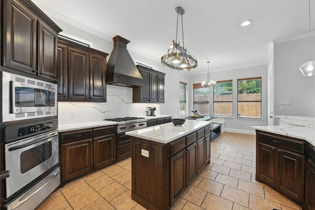 Chef's kitchen features Wolf gas cooktop, stainless steel appliances, ornamental molding, decorative light fixtures, custom exhaust hood, upgraded light granite counters and a notable pot rack. | Image 12