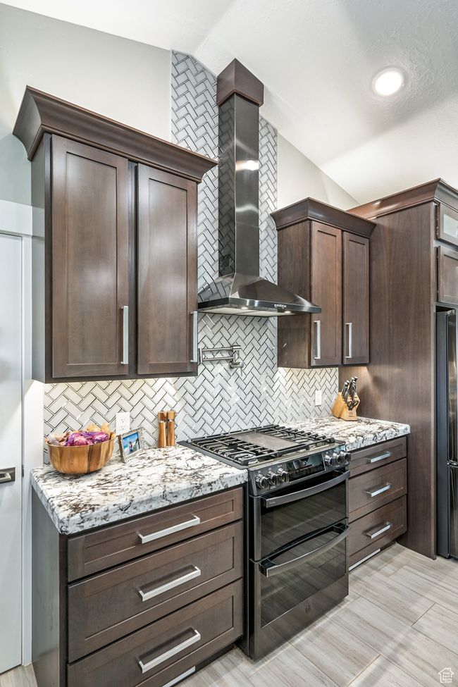 Kitchen featuring range with two ovens, wall chimney exhaust hood, vaulted ceiling, backsplash, and light stone countertops | Image 28