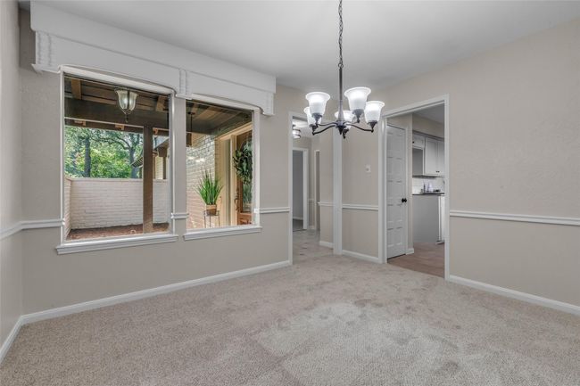 This photo showcases a cozy dining area with soft carpet flooring, neutral walls, and an elegant chandelier. Large windows provide ample natural light and a view of the courtyard, creating an inviting atmosphere. The room connects seamlessly to a kitchen, visible through an open doorway. | Image 12