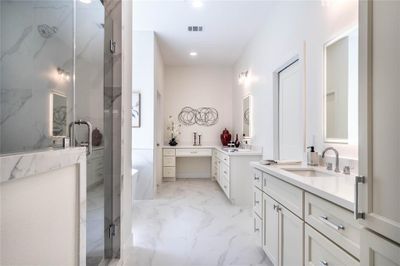 Clean-lined bathroom featuring double vanities with lots of storage, backlit mirrors, and a spacious walk-in shower with glass doors. The room is finished with sleek white cabinetry and designer tiles, giving it a modern aesthethic feel. | Image 1