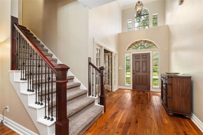 The view back toward the entry highlights the graceful staircase with wood steps and iron railings, paired with large windows that bathe the space in natural light. A classic overhead fixture brings a timeless touch to the soaring ceiling. | Image 8