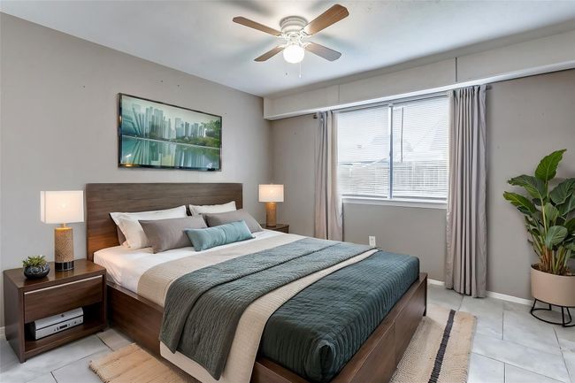 This primary bedroom boasts a serene atmosphere with its neutral palette and modern tile flooring. Natural light flows through the large window, creating a warm and inviting retreat. A ceiling fan enhances comfort, making this space a peaceful escape. | Image 11