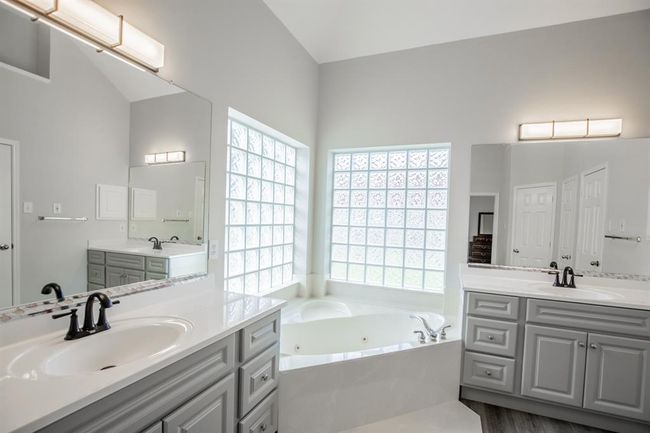 This master bathroom features a modern design with dual sinks, sleek fixtures, and ample storage. It includes a large soaking tub set beneath a glass block window, providing natural light and privacy. | Image 29