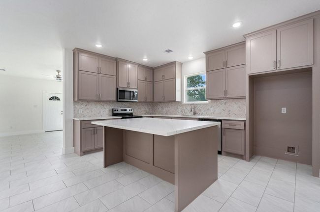 This photo shows a modern kitchen with sleek taupe cabinetry, a large central island with a marble countertop, and stainless steel appliances. The space is bright with recessed lighting and a window for natural daylight. The open layout flows into a tiled living area. | Image 15