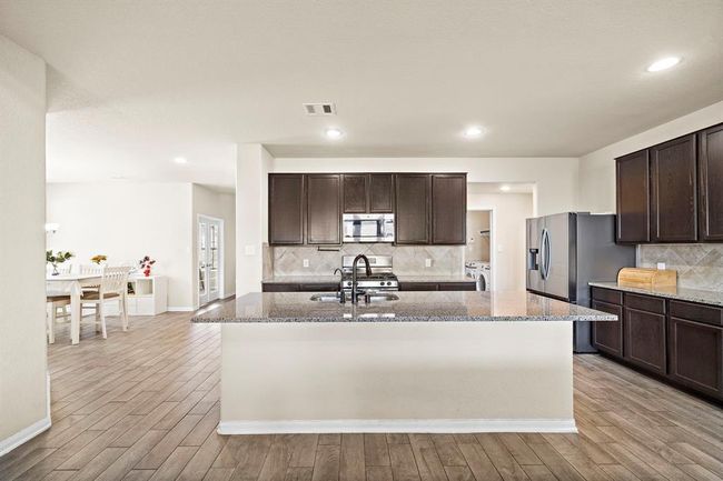 This photo showcases a spacious, modern kitchen featuring dark wood cabinetry, a large central island with a granite countertop, and stainless steel appliances. The open layout seamlessly connects to a bright dining area, creating an inviting space for entertaining. | Image 7