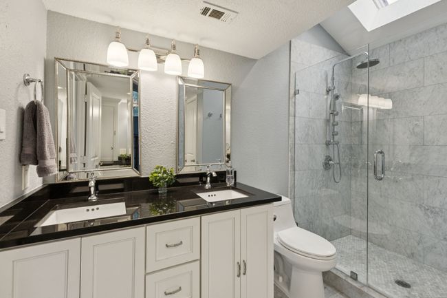 Bathroom featuring a textured wall, a stall shower, double vanity, vaulted ceiling, and toilet | Image 20
