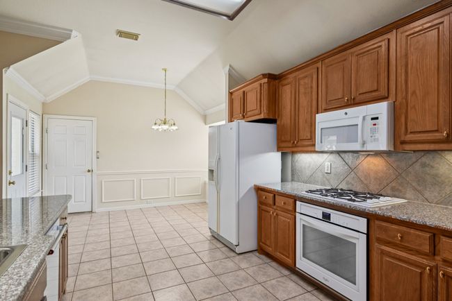 Kitchen with brown cabinetry, white appliances, light stone countertops, a wainscoted wall, and a chandelier | Image 7