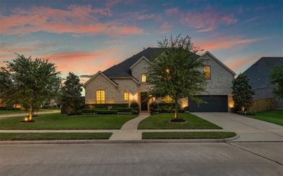 You are looking at a spacious two-story brick home with a well-manicured front lawn, featuring an attached three-car tandem garage, under the backdrop of a beautiful sunset sky. | Image 3