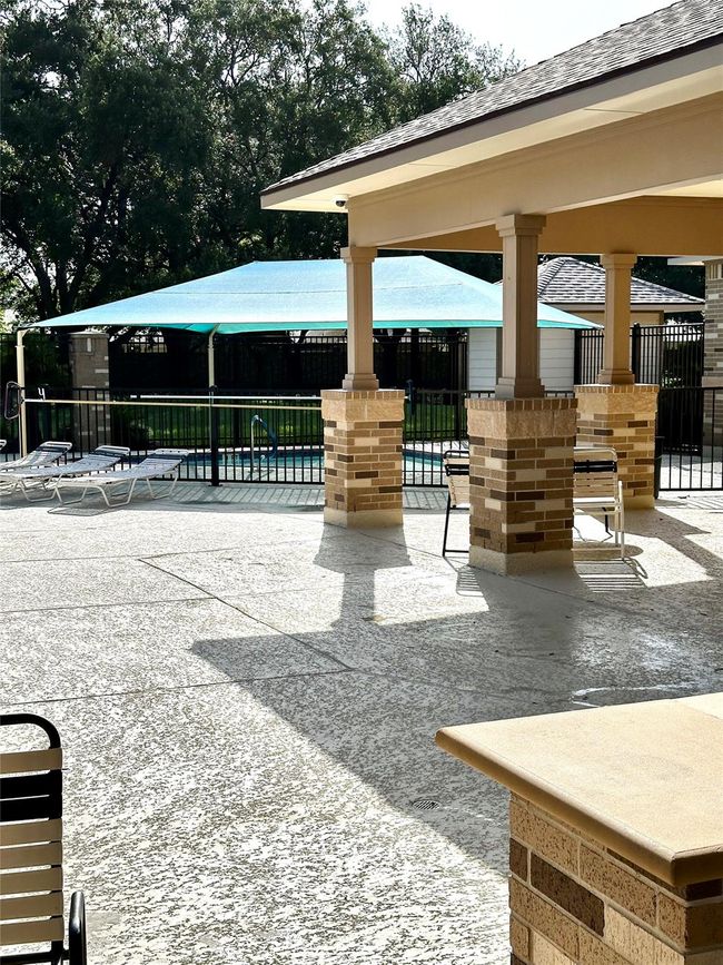 This photo shows a backyard area with a covered patio featuring brick columns, a shaded seating area, and a fenced-in pool with lounge chairs, surrounded by trees for added privacy. | Image 17