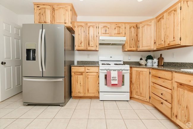 Kitchen with stainless steel fridge, white gas range, under cabinet range hood, light brown cabinets, and light tile patterned flooring | Image 59