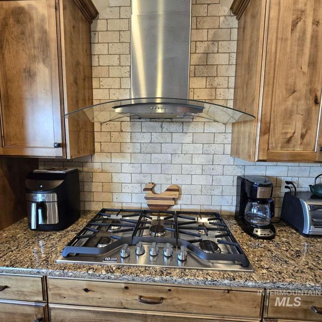 Kitchen with wall chimney exhaust hood, stainless steel gas cooktop, brown cabinetry, tasteful backsplash, and dark stone counters | Image 9