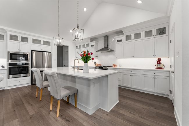 Kitchen featuring wall chimney exhaust hood, appliances with stainless steel finishes, light countertops, white cabinets, and a center island with sink | Image 5