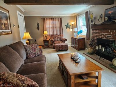 Carpeted living room featuring a baseboard heating unit, a brick fireplace, ornamental molding, and beam ceiling | Image 3