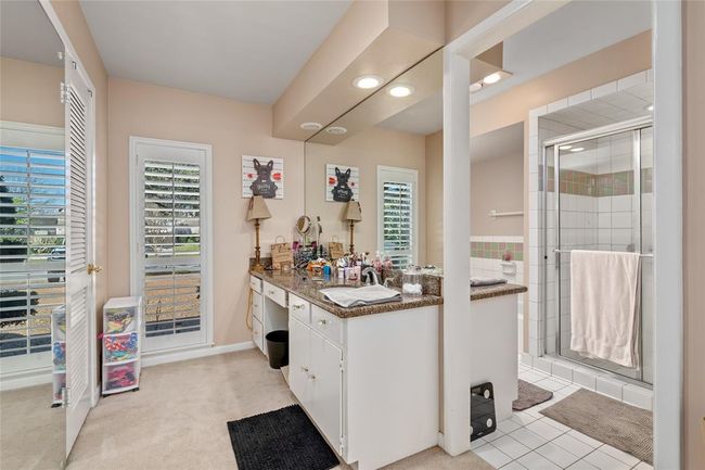The primary bathroom offers a spacious vanity with ample counter space and storage. A large mirror and built-in lighting enhance the functionality of the space. The ensuite includes a walk-in shower with a glass enclosure, accented by decorative tile. Natural light streams in through the windows with plantation shutters, creating a bright and inviting atmosphere. | Image 26