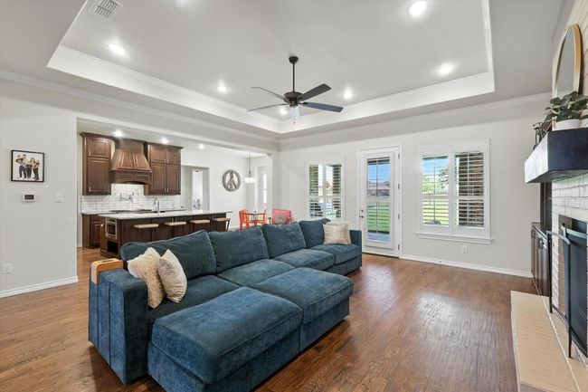 Living area featuring a tray ceiling, ceiling fan, dark wood-type flooring, crown molding, and a brick fireplace | Image 9