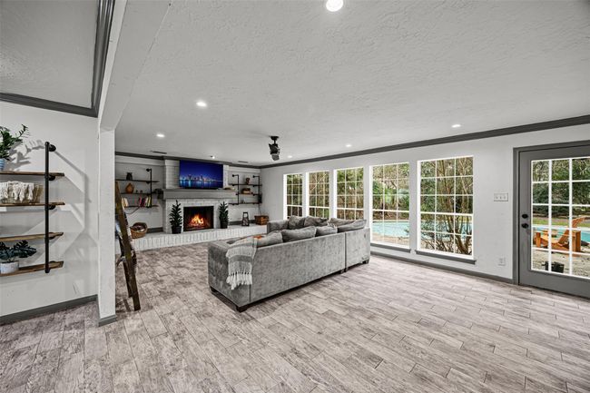 This spacious living room features large windows overlooking a pool, a cozy fireplace, modern built-in shelves, and sleek, modern tile flooring, creating an inviting and bright atmosphere. | Image 6