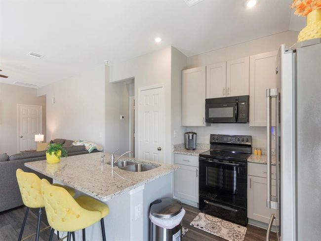 This photo showcases a modern kitchen with granite countertops, a double sink, and a breakfast bar with stylish yellow stools. It features all appliances, including a refrigerator and a range with a microwave above it. The kitchen opens into a living area with a sofa and soft lighting, creating an inviting, open-concept space. | Image 10