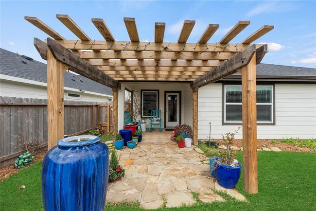 The back of the house features a door that opens up to a charming pergola with a stone patio, offering a perfect space for outdoor relaxation and entertainment. Beyond the patio, a lush green lawn stretches out, fenced in for privacy and security, creating an ideal spot for pets or outdoor activities. | Image 18