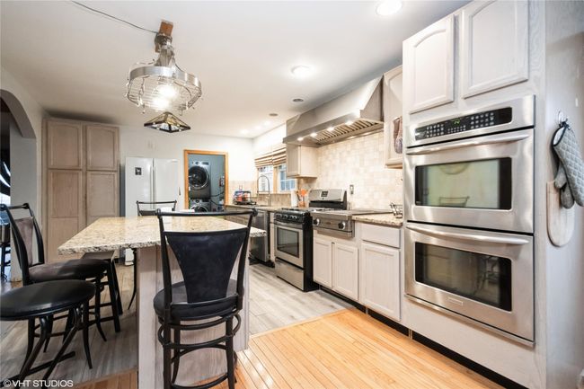 Kitchen with light stone counters, wall chimney exhaust hood, light wood-style flooring, stainless steel appliances, and decorative backsplash | Image 10