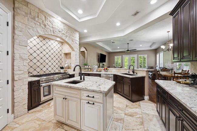 This spacious kitchen features elegant stone accents, a large island with dual sinks, and modern appliances. It opens to a bright living and dining area with hardwood floors, offering a seamless flow for entertaining. | Image 6