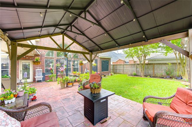 This photo showcases a cozy backyard with a covered patio featuring comfortable seating and a central fire pit. The area is surrounded by a well-maintained lawn, vibrant potted plants, and a brick exterior, offering a perfect space for outdoor relaxation and entertaining. | Image 5