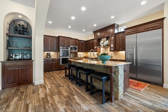 Kitchen featuring built in appliances, dark brown cabinets, backsplash, a center island with sink, and light stone counters | Image 11