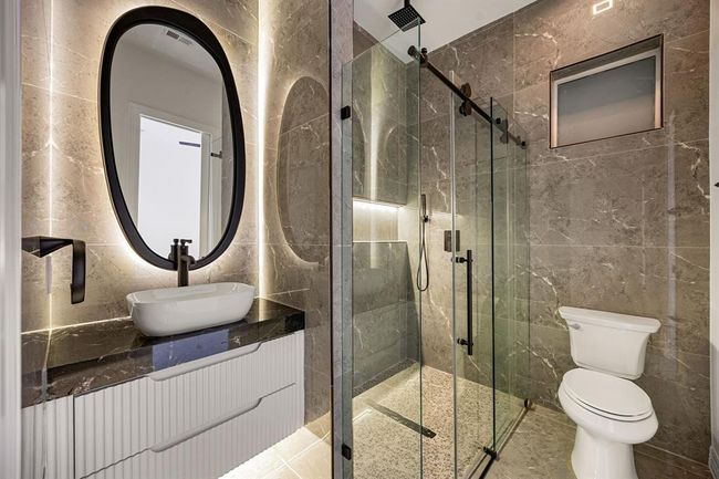 This stylish shared bathroom showcases a modern design with floor-to-ceiling stone tile, a sleek glass-enclosed shower with a rainfall showerhead, and a floating vanity topped with a vessel sink. Backlit mirrors and matte black fixtures add a touch of elegance and contemporary flair. | Image 32