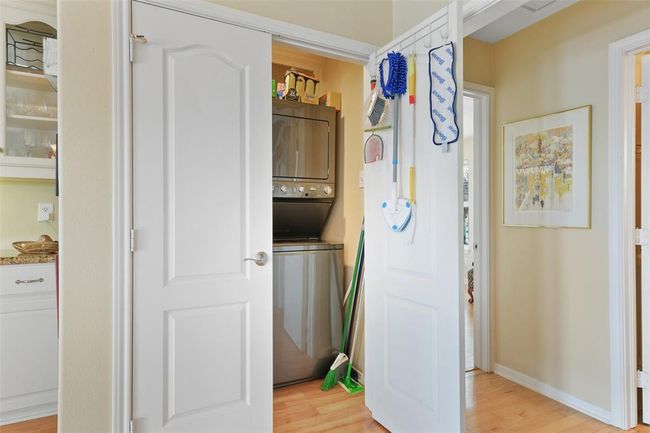 This photo shows a compact laundry area with a stacked washer and dryer, neatly tucked behind double doors. The adjacent wall has hooks for cleaning supplies. The space is conveniently located near a hallway and kitchen, featuring light-colored walls and wood flooring. | Image 21