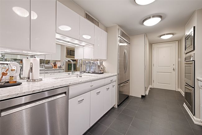 Galley kitchen featuring stainless steel appliances, white cabinetry, dark tile patterned flooring, and marble counters. Facing the back door to hallway. | Image 6