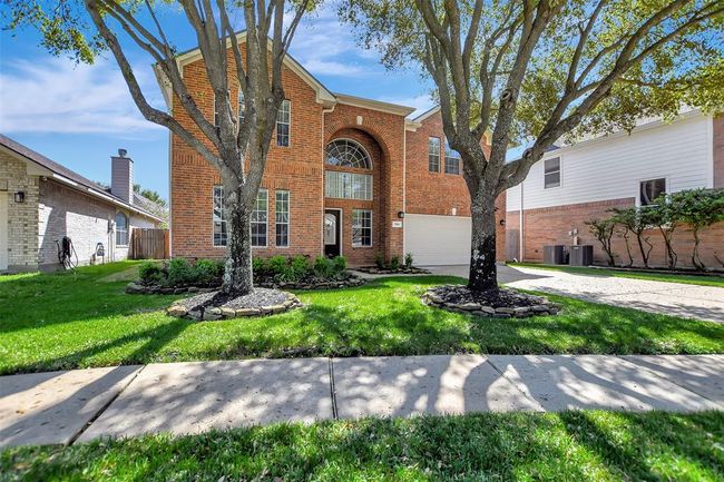 This is a two-story brick home with large windows and a two-car garage. The front yard features manicured landscaping and mature trees, offering a welcoming and shaded entrance. | Image 4