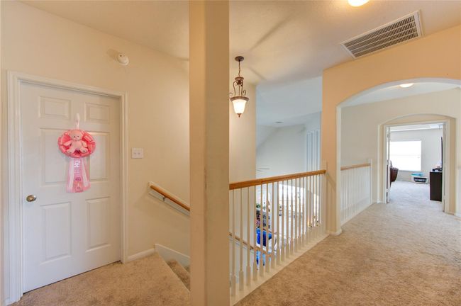 This photo shows a bright, carpeted upstairs hallway with a decorative light fixture. It features a door on the left, a staircase with a wooden handrail, and an open archway leading to another room, creating a spacious and inviting atmosphere. | Image 31