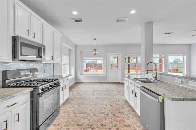 This spacious and bright kitchen features modern stainless steel appliances, white cabinetry, and a stylish tiled backsplash. The open layout includes a large island with a sink and a view of the backyard through multiple windows, enhancing the airy feel. | Image 16