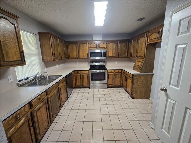 Kitchen with appliances with stainless steel finishes, light tile patterned floors, light countertops, brown cabinetry, and a textured ceiling | Image 7