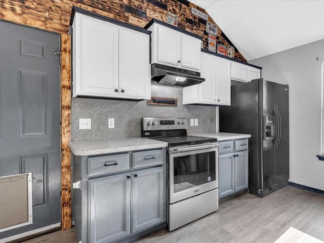 Kitchen featuring stainless steel range with electric cooktop, light wood-style floors, under cabinet range hood, vaulted ceiling, and black refrigerator with ice dispenser | Image 7