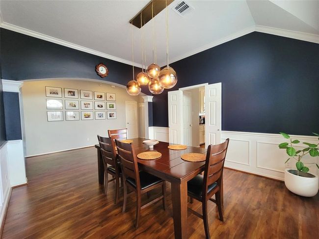 This elegant dining room features dark accent walls, wainscoting, and warm wood flooring. A modern, multi-globe chandelier hangs above a wooden dining set. The space is accented by wall art and a touch of greenery, creating a welcoming atmosphere. | Image 6
