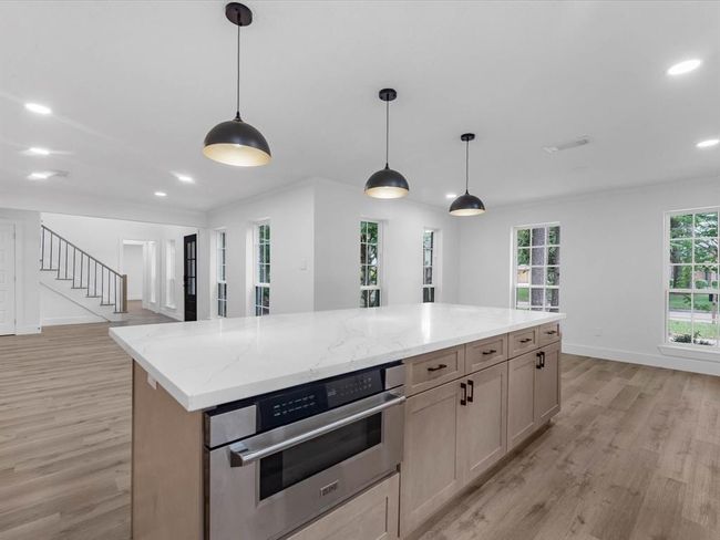 This spacious, modern kitchen features a large island with a marble countertop, pendant lighting, and ample cabinetry. The open floor plan leads to a bright living area with hardwood floors and large windows, offering a welcoming and airy atmosphere. | Image 13