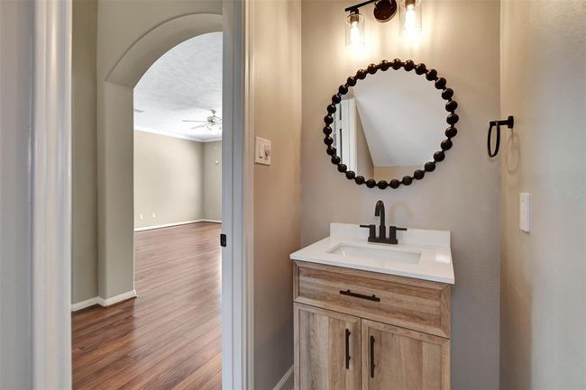 This photo features a cozy bathroom with a wooden vanity, round mirror, and modern lighting. It opens into a spacious room with wood flooring and a ceiling fan, creating a seamless transition between spaces. | Image 18