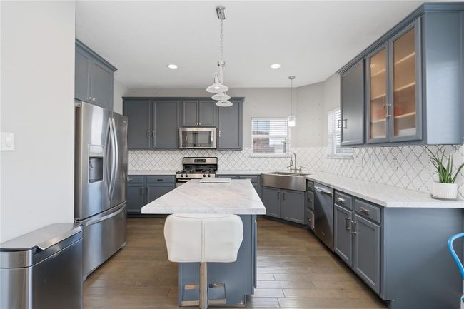 Modern kitchen with sleek gray cabinets, stainless steel appliances, and a central island with a marble countertop. The room features a farmhouse sink, stylish pendant lighting, and a geometric tile backsplash, creating a bright and inviting space. | Image 27