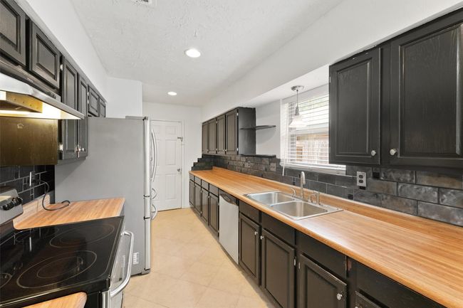This galley kitchen features sleek black cabinetry, wood countertops, and modern stainless steel appliances. The space includes a tile backsplash, dual sink, and ample natural light from a window over the sink. | Image 14