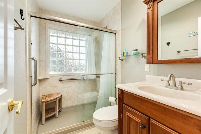 This bathroom features a glass-enclosed shower with a built-in bench and grab bars, a wooden vanity with a large mirror, and a frosted glass window for natural light. | Image 19