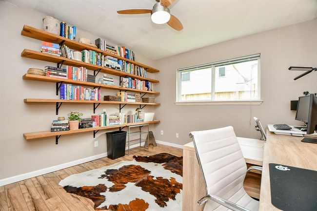 This bright home office features a large window, open shelving filled with books, a modern ceiling fan, and a stylish desk setup. The room has a warm wood floor and a cowhide rug, creating a cozy yet functional workspace. | Image 34