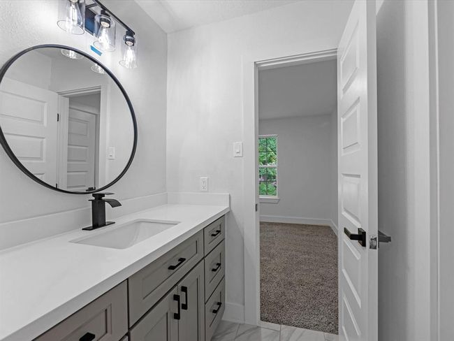 This photo shows a modern bathroom with a sleek vanity featuring a white countertop and black fixtures. A round mirror and stylish light fixtures enhance the space. A door leads to a carpeted room with a window. | Image 27