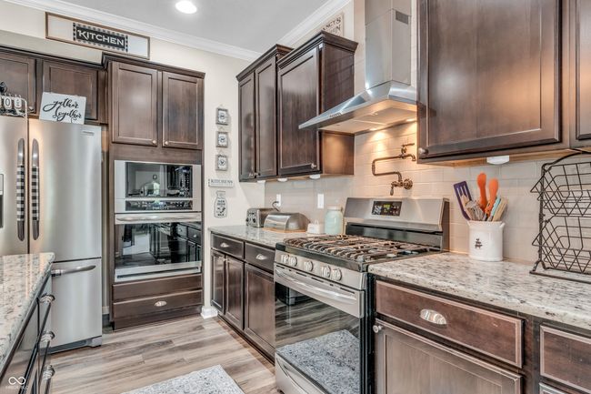kitchen features tiled backsplash, chimney vent, stainless steel appliances, ornamental molding, dark brown cabinets, and light granite counter tops | Image 31