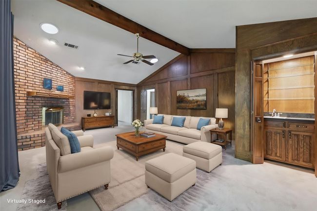 Virtually Staged Carpeted living room featuring lofted ceiling with beams, visible vents, ceiling fan, wooden walls, and a fireplace | Image 18
