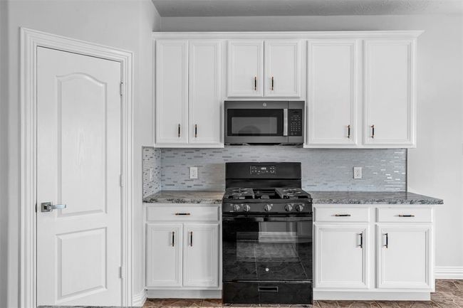 This kitchen features modern white cabinetry with sleek hardware, a stylish tile backsplash, and a black gas range with a matching microwave. The granite countertops add a touch of elegance, and the space is well-lit and functional. | Image 13
