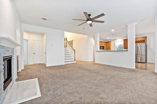 This spacious open-concept living area features a cozy carpeted floor, a modern ceiling fan, and a stylish fireplace. The kitchen boasts stainless steel appliances and warm wooden cabinetry, and the staircase adds a welcoming touch. Perfect for comfortable family living. | Image 11
