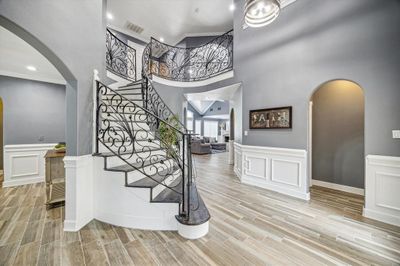 Elegant entryway with a sweeping staircase featuring ornate ironwork, complemented by modern gray tones and wood-look tile flooring that flows into a spacious, well-lit living area. | Image 1